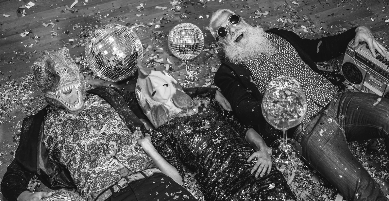 Three people in sequined outfits and quirky masks lying on the floor surrounded by confetti, one holding a boombox, at an event management company party.